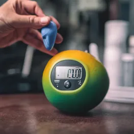 A close-up photograph of a person's hand gripping a stress ball, while a blood sugar monitor is visible in the background, emphasizing the connection between work stress and its impact on blood sugar levels.